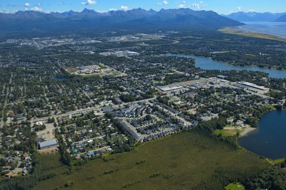 An aerial view of the Anchorage metropolitan area shows sprawling development surrounded by tall mountains.